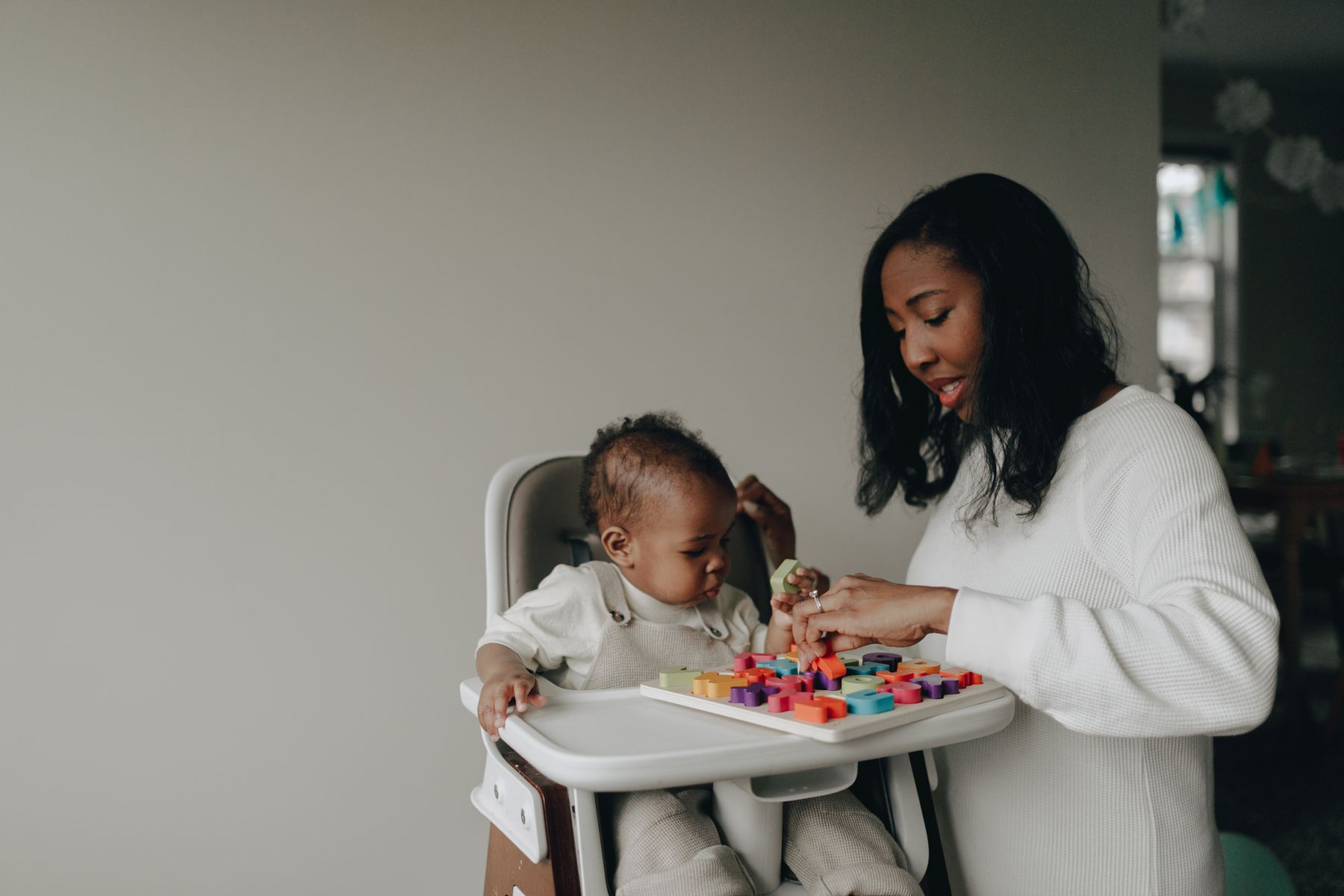 A baby in a high chair working a colourful shape puzzle with a caregiver