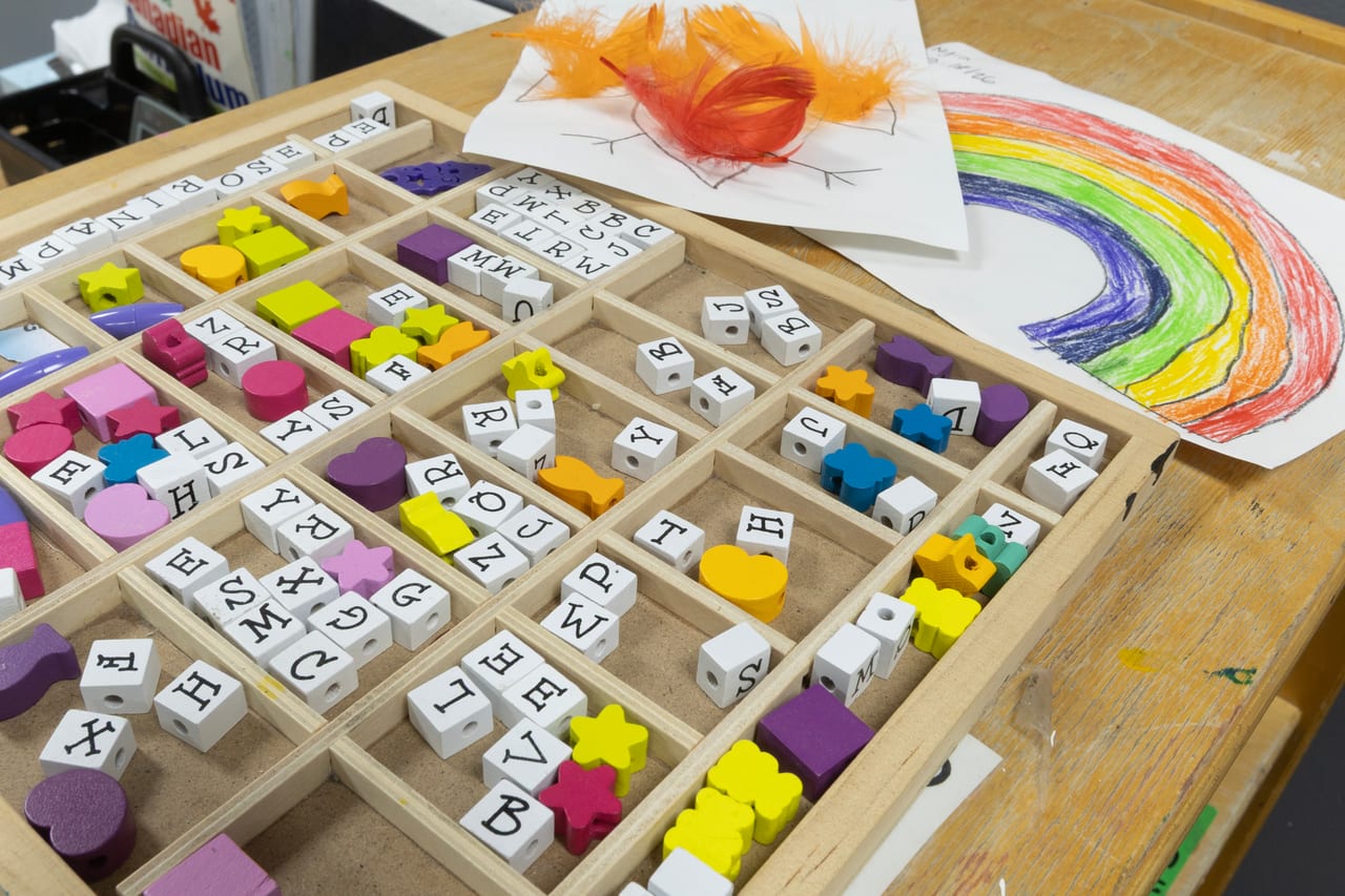 A sorting tray of alphabet and shape beads beside a child's rainbow drawing