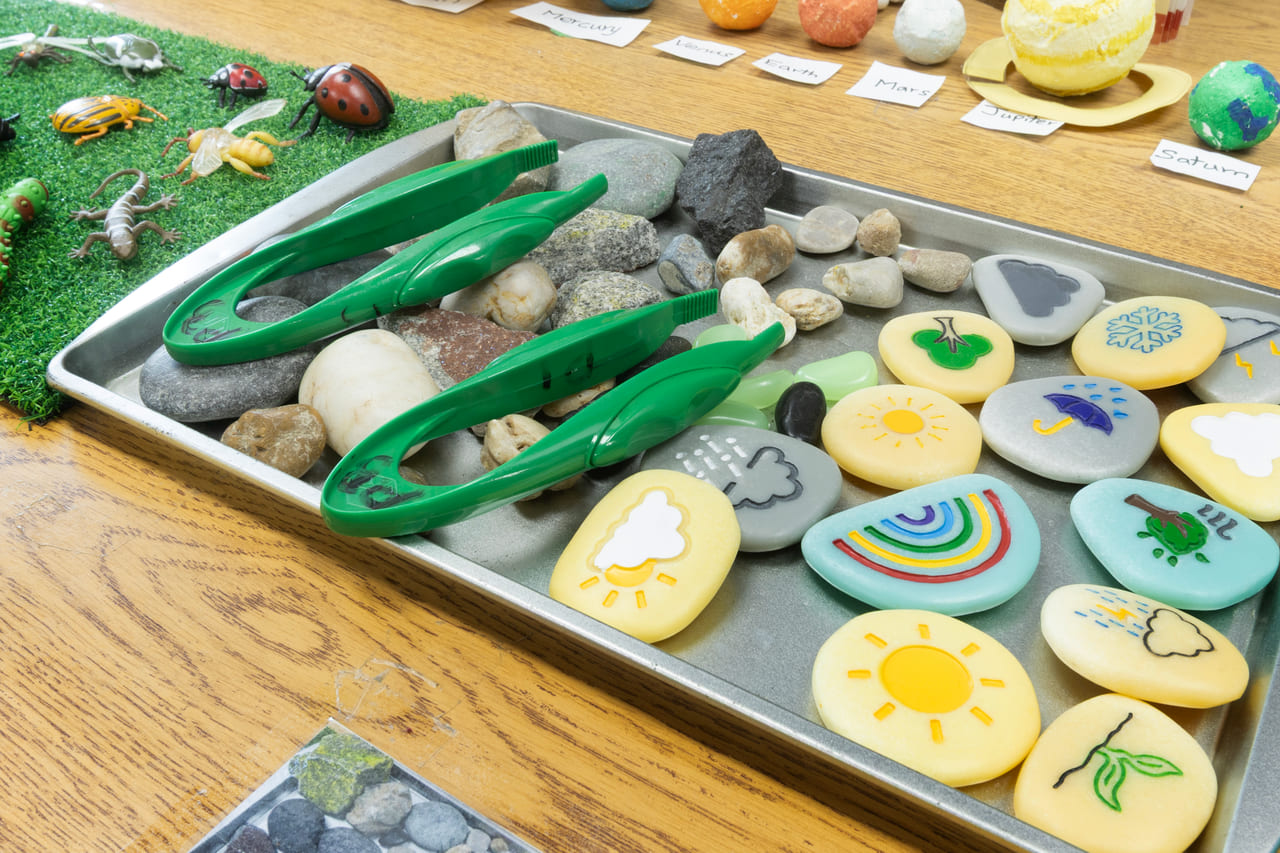 Painted story stones showing weather and nature symbols, laid out on a tray with tongs